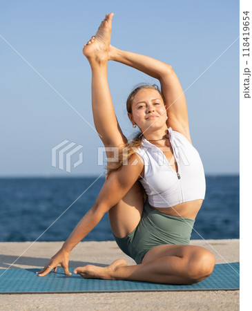 Young girl in sportwear doing stretching exercises for strong arms and abs and difficult yoga asana on the seashore on sunny day 118419654