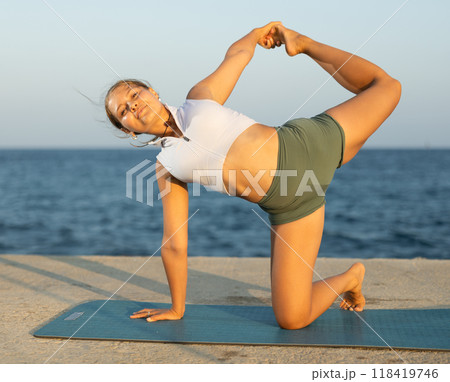 On windy day young slender woman does yoga near sea doing Crescent Moon pose 118419746
