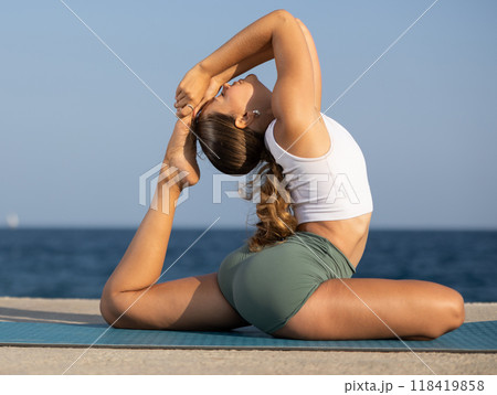 Flexible fit woman doing yoga on the seashore, performing seated back-bending asana Rajakapotasana, or King Pigeon Pose Flexible fit woman doing yoga on the seashore, performing seated back-bending asana Rajakapotasana, or King Pigeon Pose 118419858