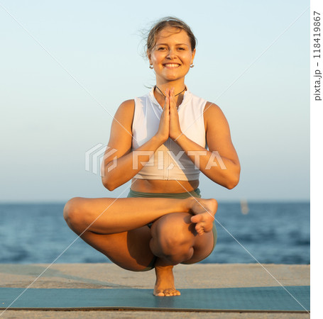 Sportive girl practicing one legged chair pose of yoga on seashore Sportive girl practicing one legged chair pose of yoga on seashore 118419867