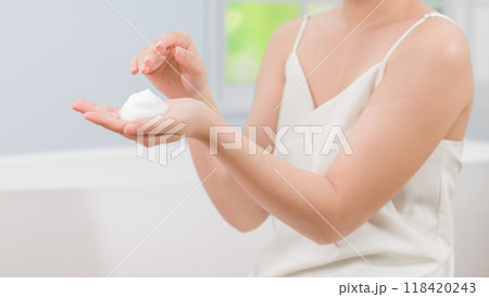 A woman holds a dollop of fluffy cream in her palm, preparing to apply it. The bright bathroom setting enhances the feeling of freshness suitable for Skincare advertising. 118420243