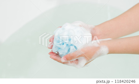 With soapy hands, a woman prepares to wash herself, holding a blue loofah on the tranquil bathwater in the bathtub. Shower gel advertisement in minimalist bathroom setting. With soapy hands, a woman prepares to wash herself, holding a blue loofah on the tranquil bathwater in the bathtub. Shower gel advertisement in minimalist bathroom setting. 118420246
