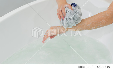 Personal care product advertisement with image hand with a loofah gently exfoliates the arm, creating a rich lather in bathtub on the minimalist space. 118420249