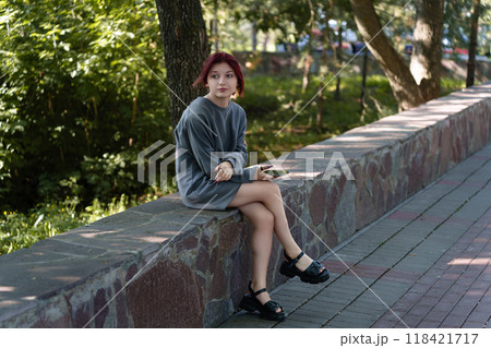 young girl with dyed hair, facial piercings and an oversize sweater holding a phone in her hand stares pensively into the distance while sitting in a summer park young girl with dyed hair, facial piercings and an oversize sweater holding a phone in her hand stares pensively into the distance while sitting in a summer park 118421717