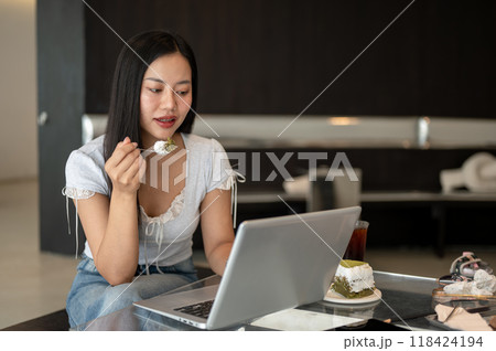 An attractive Asian woman is eating cake while focusing working on her laptop in a coffee shop. An attractive Asian woman is eating cake while focusing working on her laptop in a coffee shop. 118424194