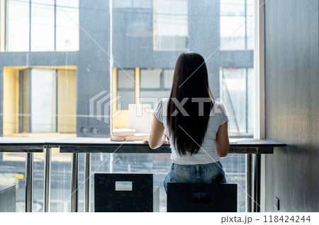 A rear view of an Asian woman sitting at a table by the window, working remotely from a coffee shop. A rear view of an Asian woman sitting at a table by the window, working remotely from a coffee shop. 118424244