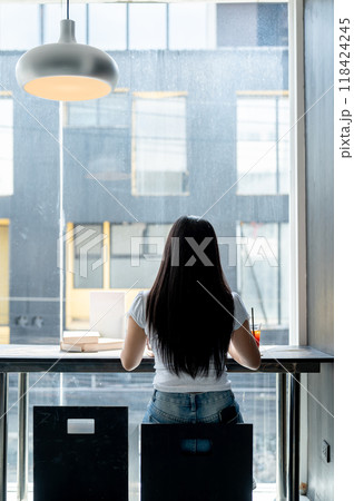 A rear view of an Asian woman sitting at a table by the window, working remotely from a coffee shop. A rear view of an Asian woman sitting at a table by the window, working remotely from a coffee shop. 118424245