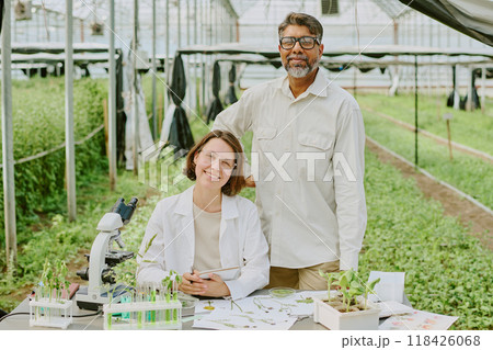 Hispanic woman and African American man standing in modern greenhouse laboratory wearing lab coats and smiling. Workspace shows laboratory equipment, plants, and research documents Hispanic woman and African American man standing in modern greenhouse laboratory wearing lab coats and smiling. Workspace shows laboratory equipment, plants, and research documents 118426068