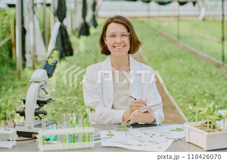 Scientist analyzing plants in laboratory greenhouse setting surrounded by equipment and samples, smiling and sitting at desk with clear test tubes, microscope, and green plants 118426070
