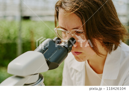 Scientist intently observing sample through microscope in laboratory setting wearing white lab coat and safety glasses, background blurred with laboratory equipment Scientist intently observing sample through microscope in laboratory setting wearing white lab coat and safety glasses, background blurred with laboratory equipment 118426104