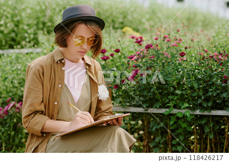Person sitting in garden and sketching in notebook while surrounded by vibrant flowers. Relaxing in casual clothing and hat, enjoying calm and serene nature environment 118426217
