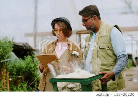 Two people discussing flower arrangements in greenhouse, standing next to lush greenery and flowers, one holding clipboard, another holding flower bouquet 118426220