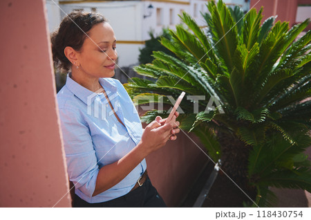 Woman in blue shirt using smartphone outdoors next to a lush green plant on a sunny day 118430754