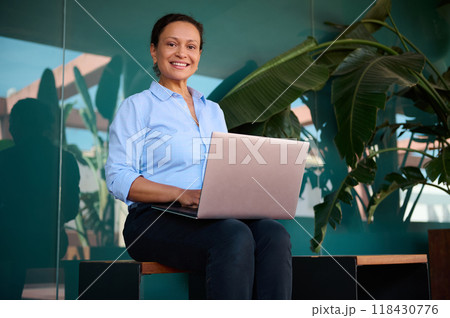 Confident professional woman smiling while working on laptop in modern outdoor setting with green plants Confident professional woman smiling while working on laptop in modern outdoor setting with green plants 118430776