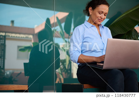Woman in casual attire working on a laptop outdoors with a reflection in glass showcasing a calm and peaceful work environment 118430777