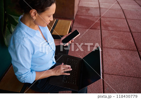 Woman in blue shirt using a laptop and smartphone on a bench outdoors for work and communication Woman in blue shirt using a laptop and smartphone on a bench outdoors for work and communication 118430779