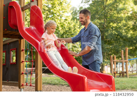 A father stands at the bottom of a red playground slide, smiling and encouraging his young daughter as she slides down. The scene takes place on a sunny summer day 118432950