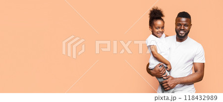 Happy black father holding his little daughter in his arms. Both are smiling at the camera and appear to be enjoying spending time together. The background is a soft peach color, copy space 118432989