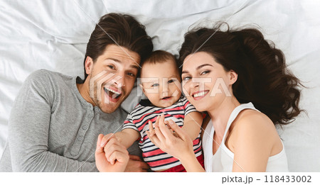 A happy family of three, a man, a woman, and their baby, are lying on a white bed with their heads together. They are all smiling and look very happy, top view 118433002