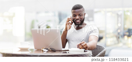 A man with a beard sits at a table outdoors, talking on a phone while working on a laptop and taking notes. A man with a beard sits at a table outdoors, talking on a phone while working on a laptop and taking notes. 118433021