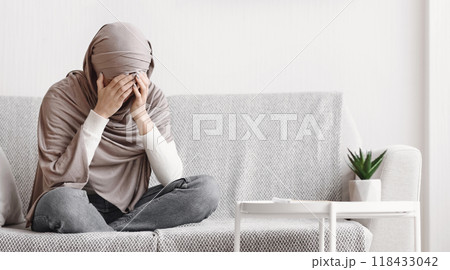 Depression concept. Sad muslim woman in hijab crying on couch at home, covering her face with hands, free space 118433042