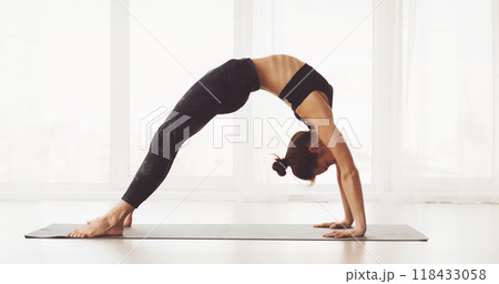 A woman is showcased in a spacious, well-lit studio, expertly executing a back-bending yoga pose. She balances on her palms and feet, with her back arched and hips elevated A woman is showcased in a spacious, well-lit studio, expertly executing a back-bending yoga pose. She balances on her palms and feet, with her back arched and hips elevated 118433058
