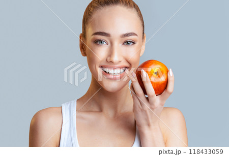 A woman with blonde hair and bright blue eyes smiles broadly as she holds a red apple in her hand. The background is a plain grey, allowing her bright smile and the vibrant apple to stand out. 118433059
