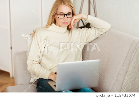 A woman sits on a couch in her living room, wearing glasses and a white sweater, working on a laptop computer. 118433074