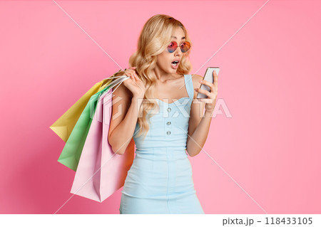A young woman with long blonde hair is standing in a pink studio, wearing sunglasses and a blue dress. She is holding a smartphone in her right hand, her eyes widened in surprise. A young woman with long blonde hair is standing in a pink studio, wearing sunglasses and a blue dress. She is holding a smartphone in her right hand, her eyes widened in surprise. 118433105