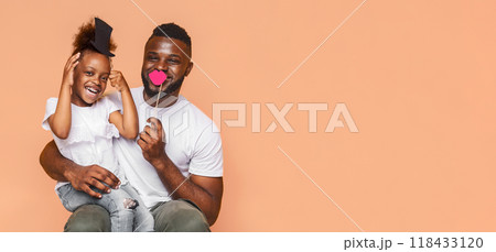 Cute black father and daughter are sitting together on an orange background. The little girl is wearing a black hat and holding up a pink heart while the father is holding up a pink mustache. 118433120