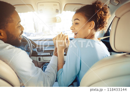 African American couple is sitting in the front seats of a car. The woman is looking over her shoulder with her mouth open in excitement and the man is looking forward, holding her hand African American couple is sitting in the front seats of a car. The woman is looking over her shoulder with her mouth open in excitement and the man is looking forward, holding her hand 118433144
