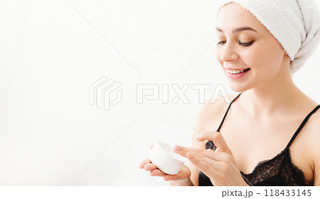 A cheerful young woman with a towel wrapped around her head is applying a creamy skincare product to her face. She is indoors, in a well-lit setting, and appears to enjoy her morning beauty routine. 118433145