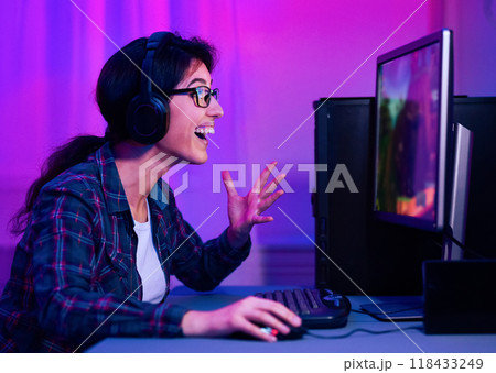 The image shows a woman wearing glasses and headphones, smiling and excitedly playing video games on her computer late at night. She has her hands on the keyboard and mouse 118433249