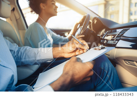 A driving instructor sits in the passenger seat of a car, holding a clipboard and pen, evaluating a student who is behind the wheel. Instructor appears to be taking notes on the students performance A driving instructor sits in the passenger seat of a car, holding a clipboard and pen, evaluating a student who is behind the wheel. Instructor appears to be taking notes on the students performance 118433343