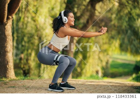 A woman is doing squats in a park, wearing a white tank top, gray leggings, black sneakers, and a resistance band around her thighs. She has curly hair and is smiling, wearing headphones. 118433361