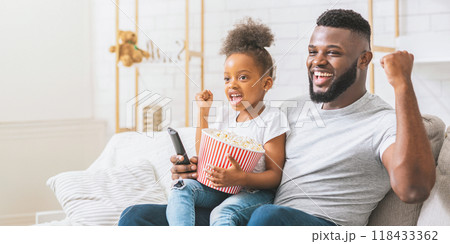 Black father and his young daughter are sitting on a couch and watching television together. They both have their hands raised in the air and look excited about what they are watching. 118433362