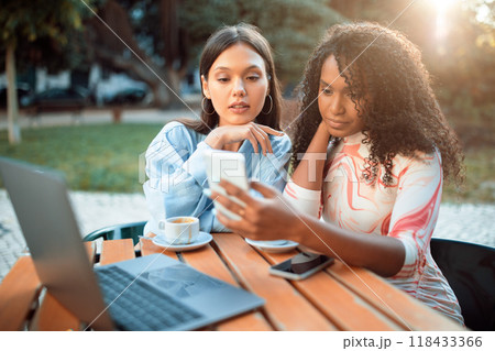 Two women sit at an outdoor cafe table made of wood. The woman with curly hair is holding a smartphone and looking at the screen, while the other woman looks on intently. Two women sit at an outdoor cafe table made of wood. The woman with curly hair is holding a smartphone and looking at the screen, while the other woman looks on intently. 118433366