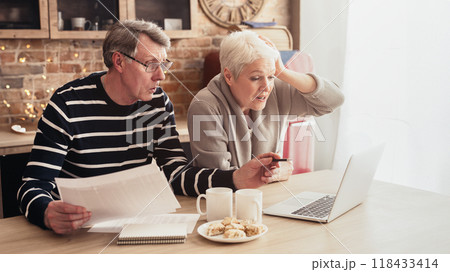 This image shows a senior couple sitting at a kitchen table, reviewing bills and a laptop together. They both have concerned expressions on faces, suggesting they are facing a financial challenge This image shows a senior couple sitting at a kitchen table, reviewing bills and a laptop together. They both have concerned expressions on faces, suggesting they are facing a financial challenge 118433414