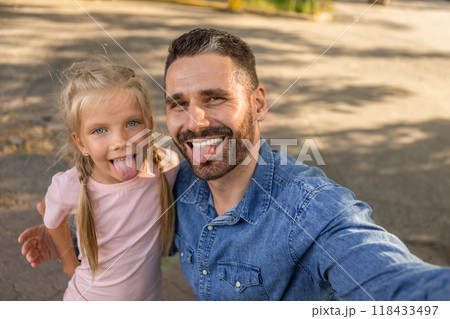 A father and his daughter are taking a selfie together outdoors. They are both smiling and sticking their tongues out playfully. They appear to be enjoying their time together. 118433497