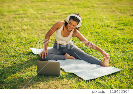 A woman with headphones is stretching her leg on a yoga mat outdoors in a park, while using a laptop. She is wearing a white sports bra and gray yoga pants. She is smiling while looking at the laptop. 118433504