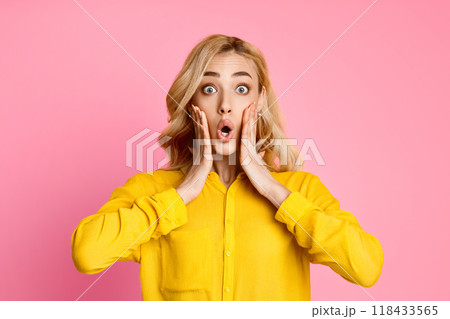 This image shows a woman in a yellow shirt with a surprised expression on her face. Her hands are placed on her cheeks, and her eyes are wide open. The background is a solid pink. This image shows a woman in a yellow shirt with a surprised expression on her face. Her hands are placed on her cheeks, and her eyes are wide open. The background is a solid pink. 118433565