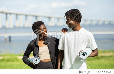 Two black people, a man and a woman, are walking together in the direction of a large bridge, each carrying a rolled-up yoga mat. They are both smiling and appear to be enjoying their time together. Two black people, a man and a woman, are walking together in the direction of a large bridge, each carrying a rolled-up yoga mat. They are both smiling and appear to be enjoying their time together. 118433598