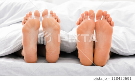 This image shows two pairs of bare feet sticking out from under white bed sheets. The feet are positioned close together, suggesting that the couple is in bed together. 118433691