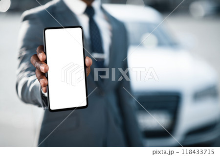 A man in a suit stands in front of a white car, holding out a smartphone with a blank white screen towards the camera, cropped 118433715
