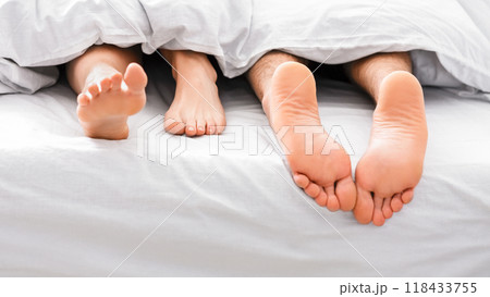 A close-up image of two bare feet, one male and one female, sticking out from underneath white bedding, suggesting a couple is sleeping in bed. A close-up image of two bare feet, one male and one female, sticking out from underneath white bedding, suggesting a couple is sleeping in bed. 118433755