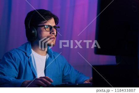 A young man wearing glasses and headphones sits at a computer in a room with purple lighting. He appears focused on the screen and may be playing a game or working on a project. A young man wearing glasses and headphones sits at a computer in a room with purple lighting. He appears focused on the screen and may be playing a game or working on a project. 118433807