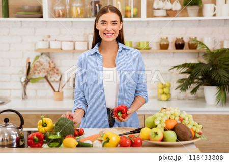 A woman smiles as she holds a red bell pepper in a modern kitchen setting, surrounded by fresh fruits and vegetables. 118433808
