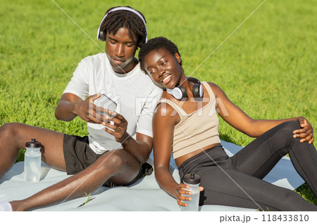African American young couple is relaxing on a blanket in a park on a sunny day. They are both wearing headphones, and the man is looking at his phone. African American young couple is relaxing on a blanket in a park on a sunny day. They are both wearing headphones, and the man is looking at his phone. 118433810