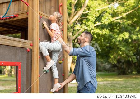 A father smiles and encourages his daughter as she climbs a wall on a playground. The young girl, with blonde hair in braids, is wearing jeans and a pink shirt and has climbed halfway up the wall. A father smiles and encourages his daughter as she climbs a wall on a playground. The young girl, with blonde hair in braids, is wearing jeans and a pink shirt and has climbed halfway up the wall. 118433849