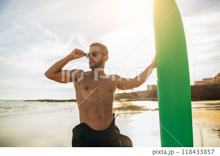 A man with a beard, wearing sunglasses, stands on a sandy beach with a green surfboard under his arm. He is shirtless and wearing black wetsuit pants. The ocean and a distant shoreline are visible 118433857